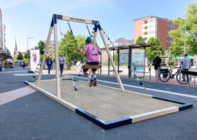 Pop-up Schaukel in der Holtenauer Straße in Kiel neben einer Bushaltestelle it Mann in rosa Shirt, der schaukelt.