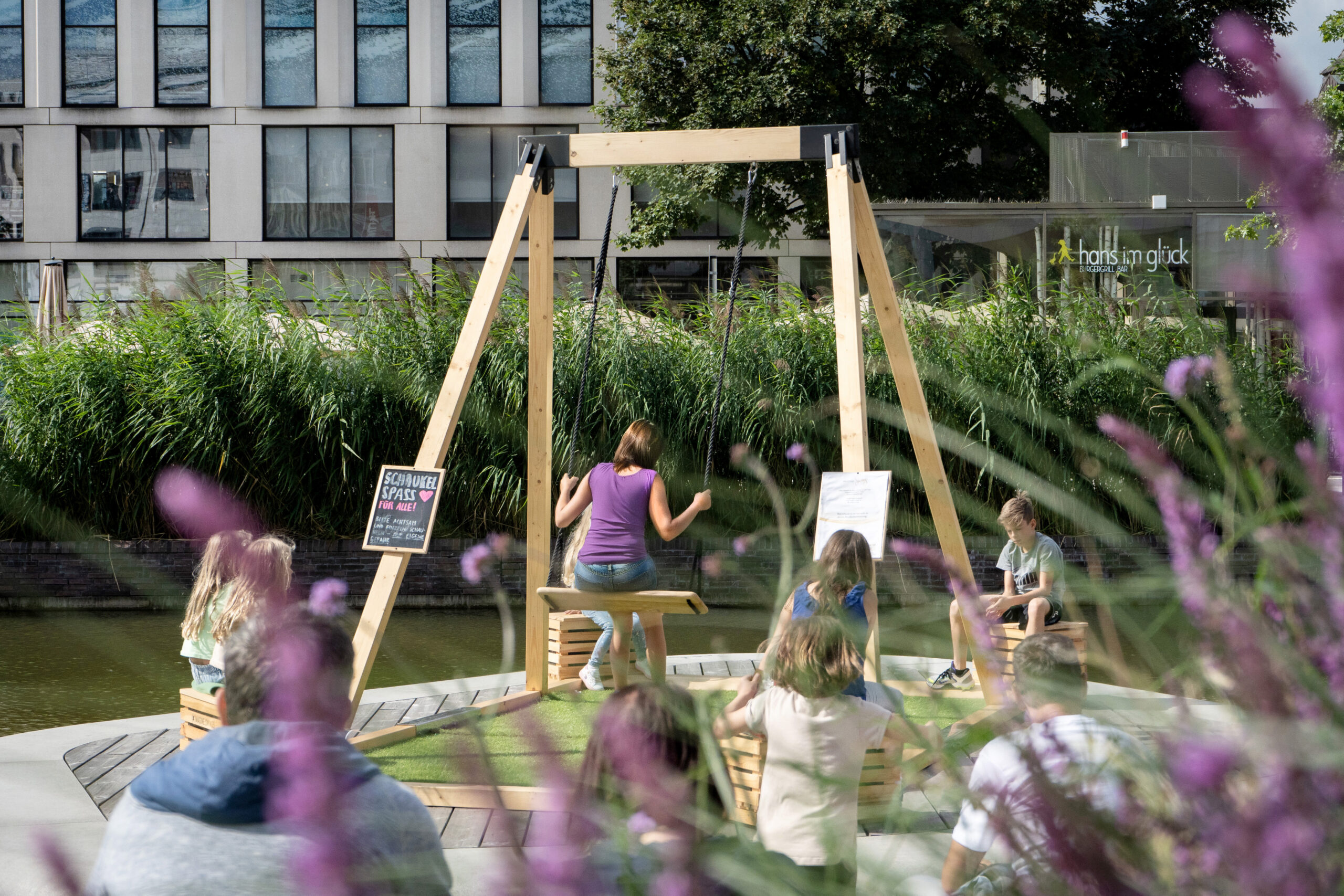Imagine-Swings_Pop-up-Schaukel_Kiel-Innenstadt-2 Mobile Pop-up Schaukel an begrüntem Ort in der Stadt mit schaukelnder Frau und Kindern rundherum