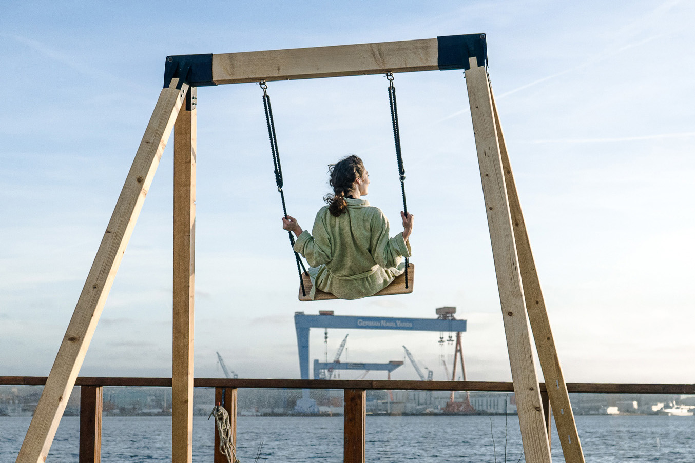Imagine-Swings_Pop-up-Schaukel_Rune-Sauna_Sandhafen_Förde-Werftblick-2 Frau schaukelt an der Förde mit Werftblick